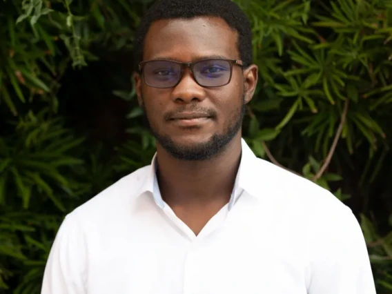 Kofi Owusu-Ansah stands against a background of leaves wearing a white shirt and glasses