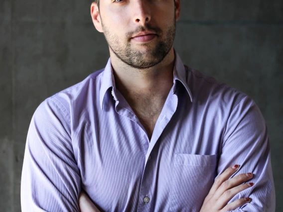 Professional photo of Lucas Belury standing in front of a matte background wearing a collared buttondown shirt with his arms crossed