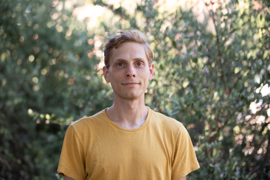 Photo of Andrew Slemmon wearing a yellow t-shirt against a background of sun-dappled leaves