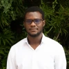 Kofi Owusu-Ansah stands against a background of leaves wearing a white shirt and glasses